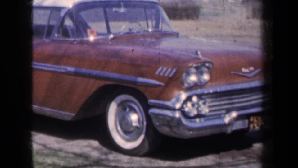 A brown Chevrolet Bel Air convertible sits on a quiet road with rural New York hills and bare trees in the background.