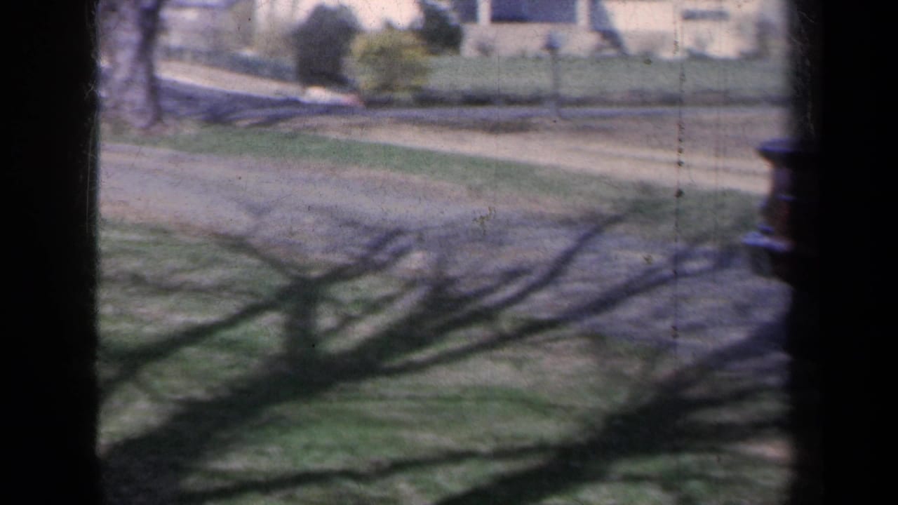 1959 New York home-movie frame looking across a suburban lawn, driveway, sidewalk, and neighboring houses in bright daylight.