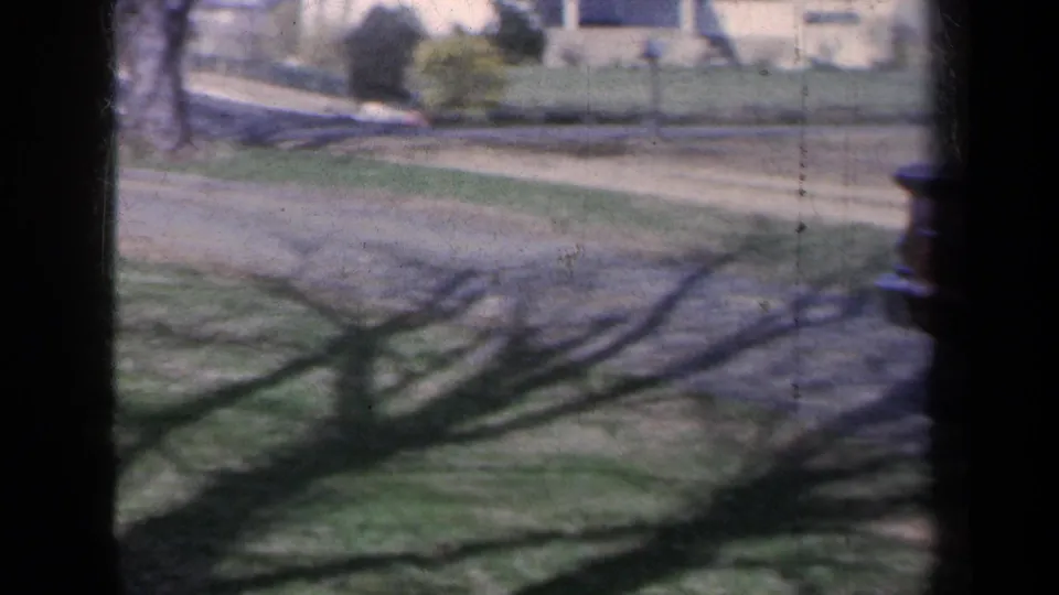 1959 New York home-movie frame looking across a suburban lawn, driveway, sidewalk, and neighboring houses in bright daylight.