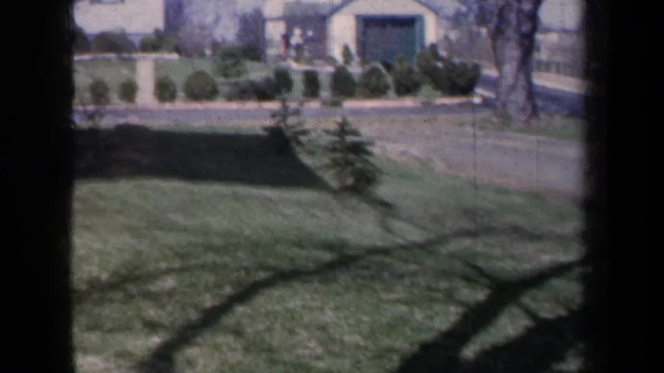 1959 New York home-movie frame showing a broad suburban yard with trimmed grass, sidewalk, shrubs, trees, and houses in the background.