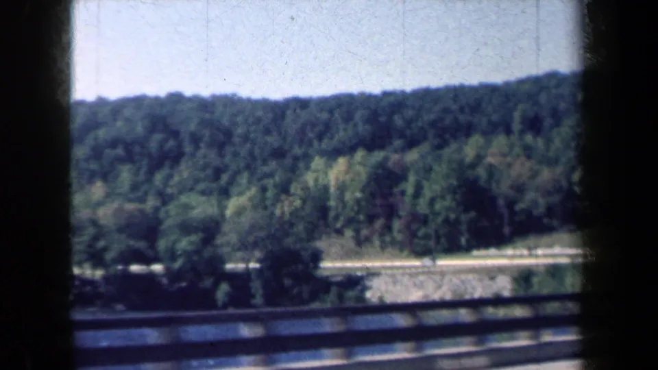 1959 New York 8mm frame with wooded hills, a roadside guardrail, and a distant road below the tree line.