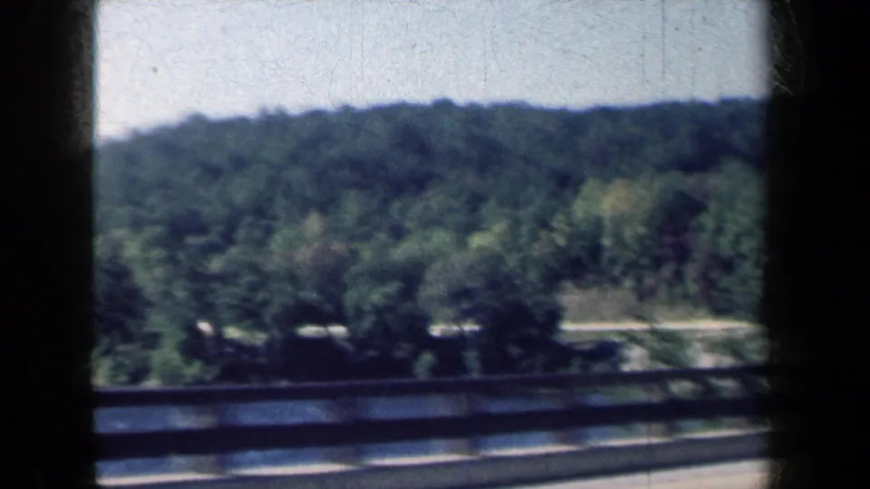 1959 New York home-movie frame showing a guardrail, tree-covered hills, and roadside motion from inside a car.