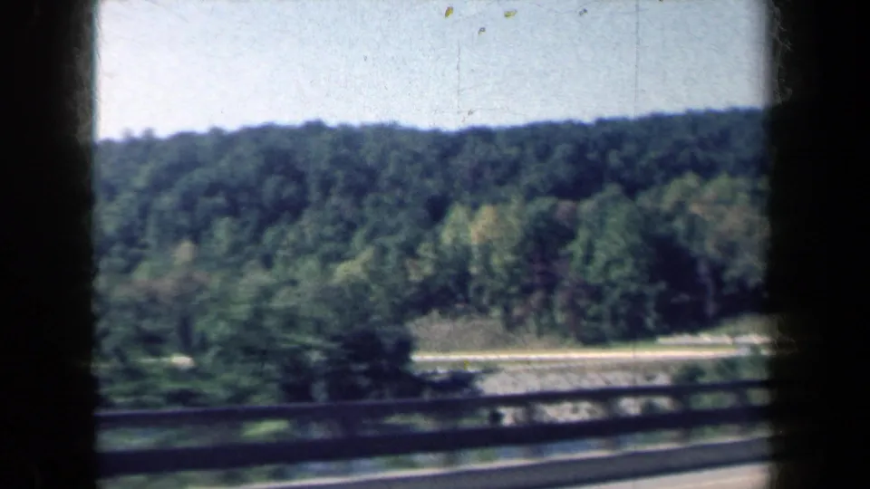 1959 8mm road-trip view of wooded New York countryside beyond a roadside guardrail and paved lane.