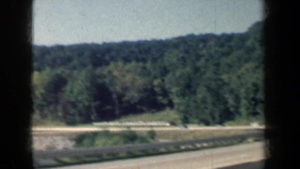 Soft 8mm view of a wooded New York highway roadside, with guardrail and pavement passing in the foreground.