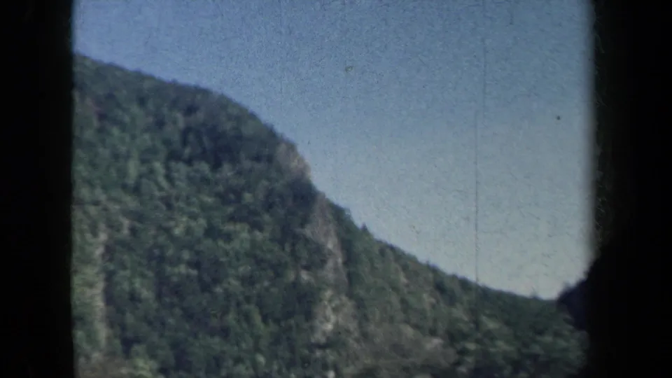 1959 8mm home-movie view of wooded mountain slopes in New York, framed by dark vehicle-window edges and soft film grain.