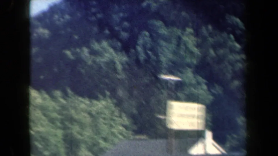 A pale roadside sign passes in front of a wooded New York hillside, softened by car motion and the 1959 8mm film texture.