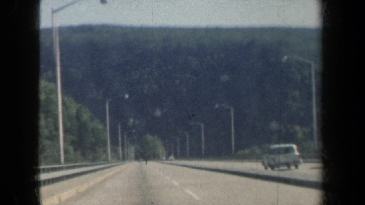Highway lanes with wooded hills, light poles, and a car at right.
