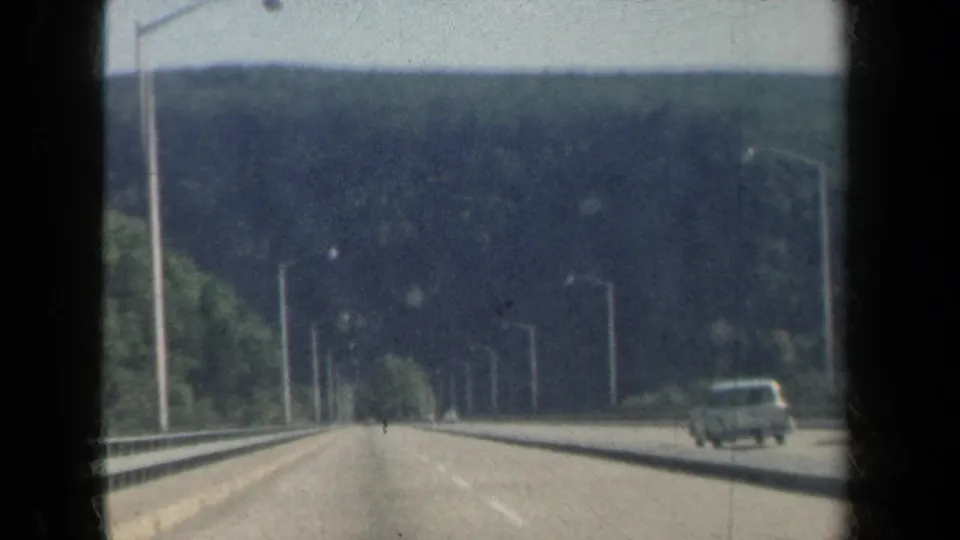 Moving 8mm freeway view in New York with forested hills, roadside lighting, and a passing car at the right edge.