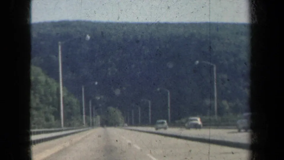 1959 New York highway footage showing multiple cars, guardrails, tall lights, and a tree-covered hill line ahead.