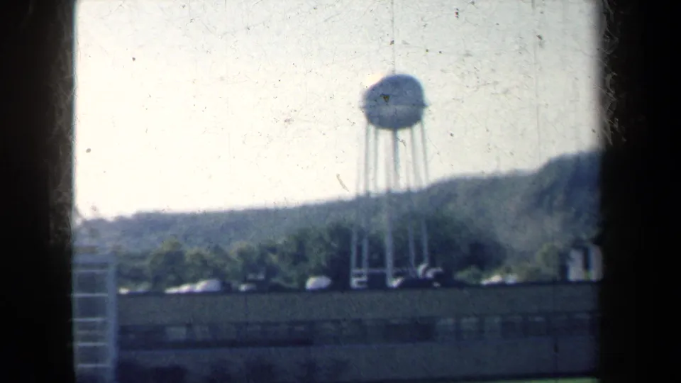 8mm New York travel view with a distant water tower, low roadside structures, parked vehicles, and wooded hills.