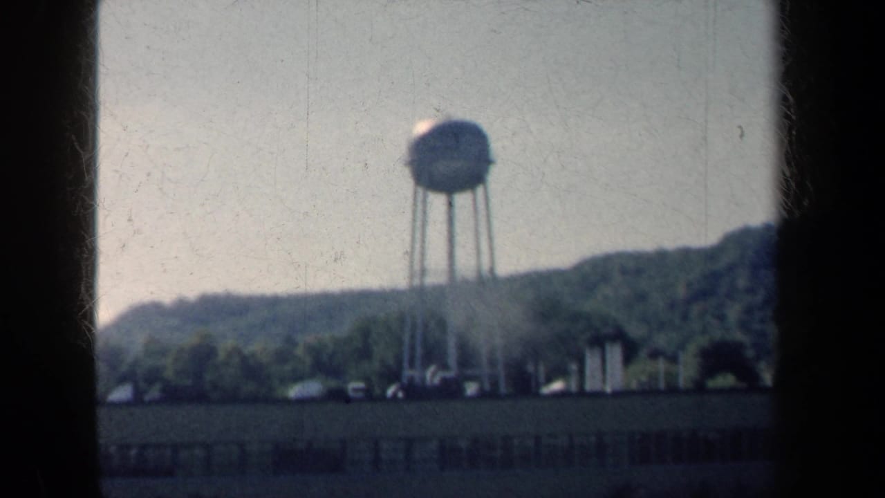 1959 New York 8mm frame showing a water tower, roadside foreground and wooded hills under a pale sky.
