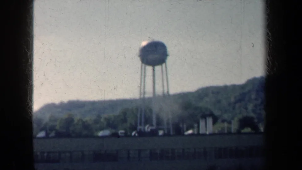 1959 New York 8mm frame showing a water tower, roadside foreground and wooded hills under a pale sky.