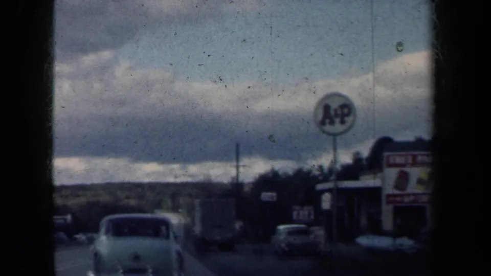 1959 New York roadside view from a moving vehicle, with a mid-century car, A&P sign and low commercial buildings.