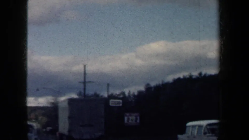 1959 New York moving-vehicle frame with roadside structures, utility poles, a parked car and distant hills.