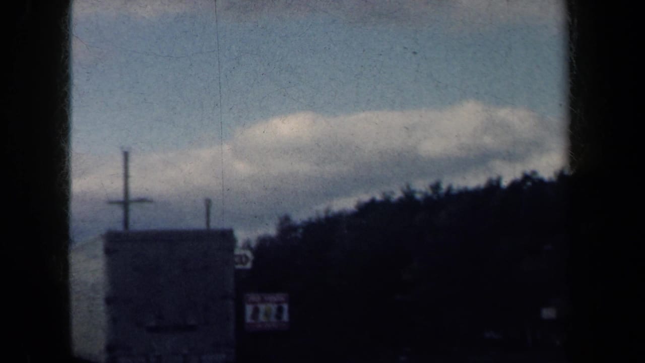 1959 New York 8mm road-trip view showing a roadside sign, tree line, cloudy sky, and dark window-frame edges from a moving vehicle.