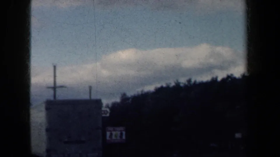 1959 New York 8mm road-trip view showing a roadside sign, tree line, cloudy sky, and dark window-frame edges from a moving vehicle.