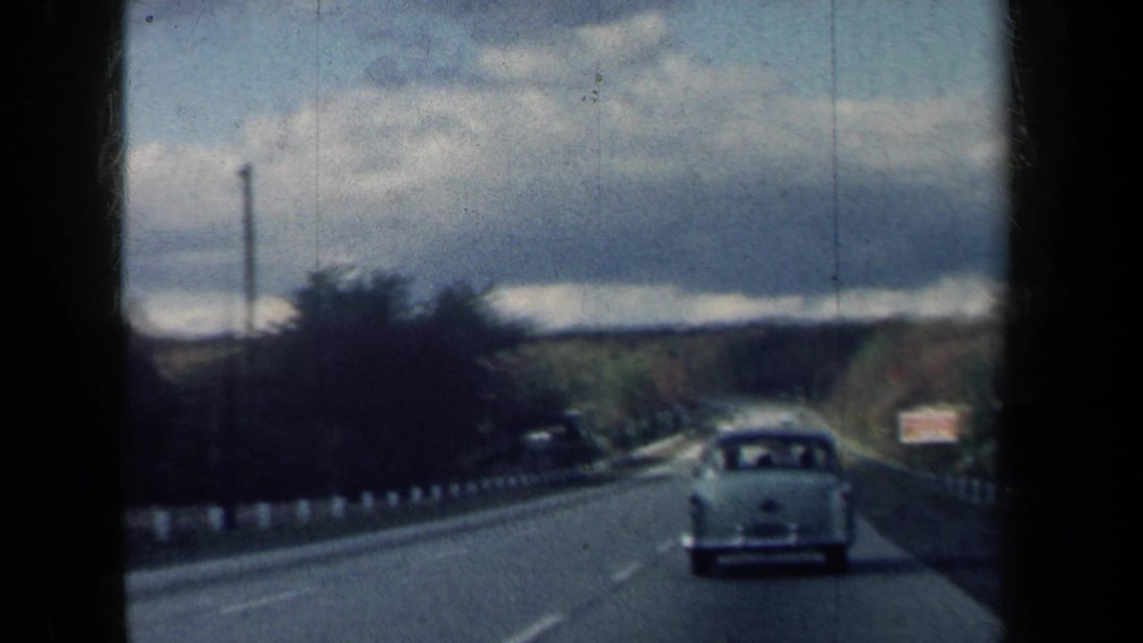 1959 New York road footage shows a white sedan ahead on a tree-lined two-lane highway with cloudy sky and distant traffic.