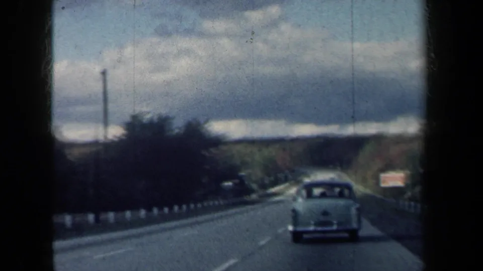 1959 New York road footage shows a white sedan ahead on a tree-lined two-lane highway with cloudy sky and distant traffic.