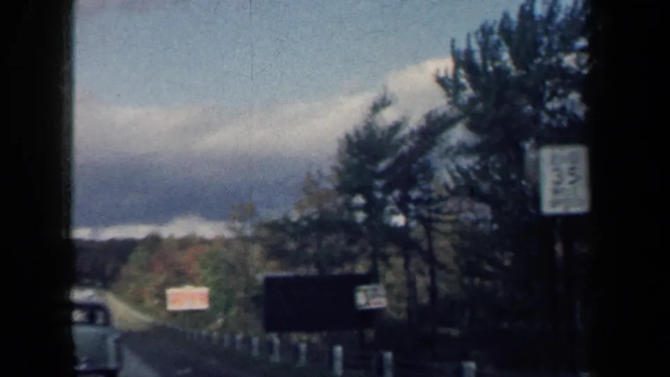 1959 New York 8mm frame shows a moving highway view with a white sedan, roadside signs, trees, and a dark vehicle window edge.