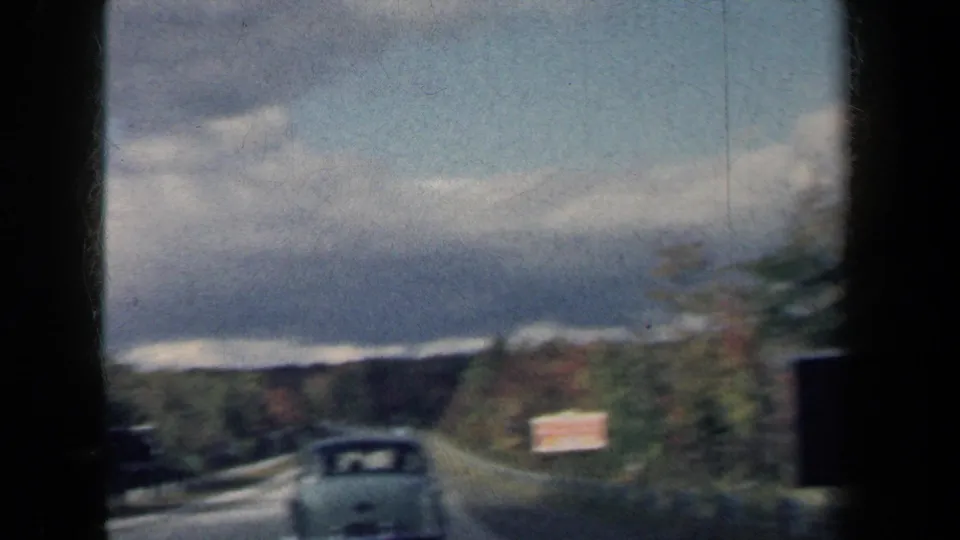 1959 New York highway view shows a white sedan approaching a curve, with a roadside sign, wooded hills, and cloudy sky.