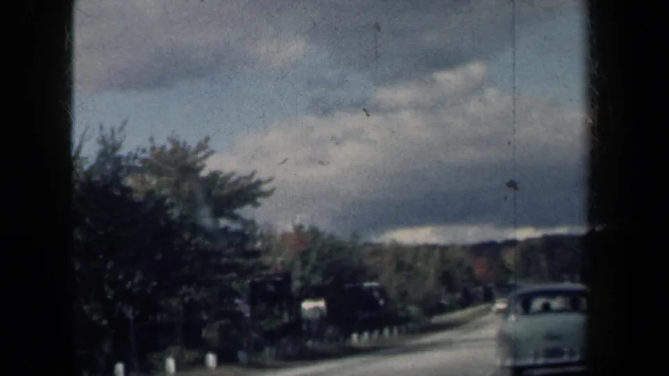 1959 New York 8mm home-movie frame showing a pale classic car ahead on a wooded road under heavy clouds.