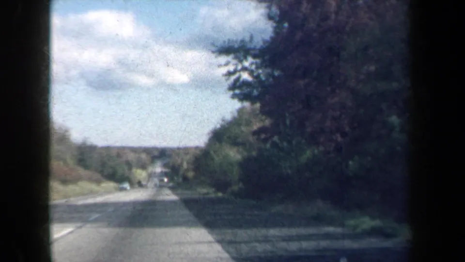 1959 New York 8mm view from a moving vehicle on a paved road lined with dense trees and distant traffic.