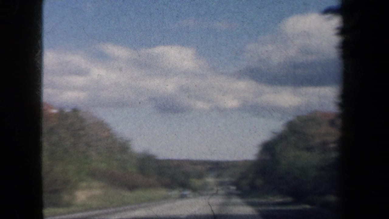 Vintage 8mm driving view along a wooded New York road with low hills, trees, and partly cloudy sky.