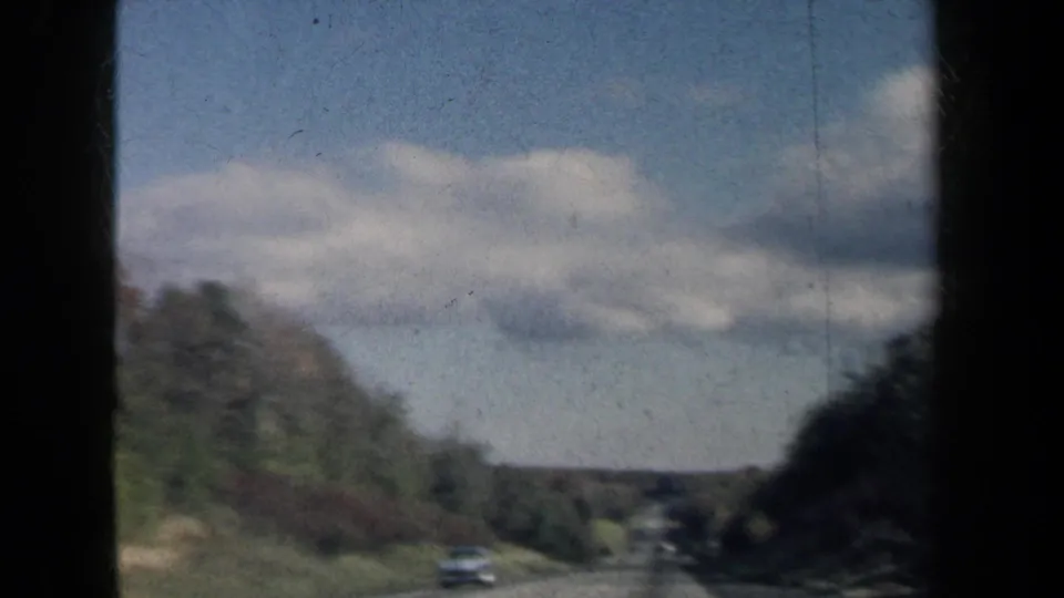1959 New York home-movie frame showing a distant road, wooded shoulders, low hills, and soft clouds from a vehicle.