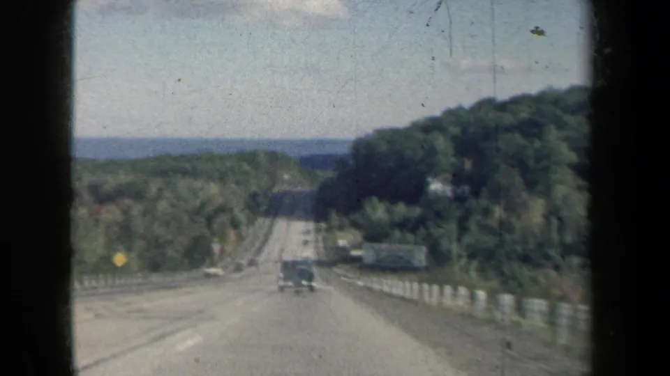 1959 New York 8mm frame showing a car-window view down a wooded highway toward distant blue water.