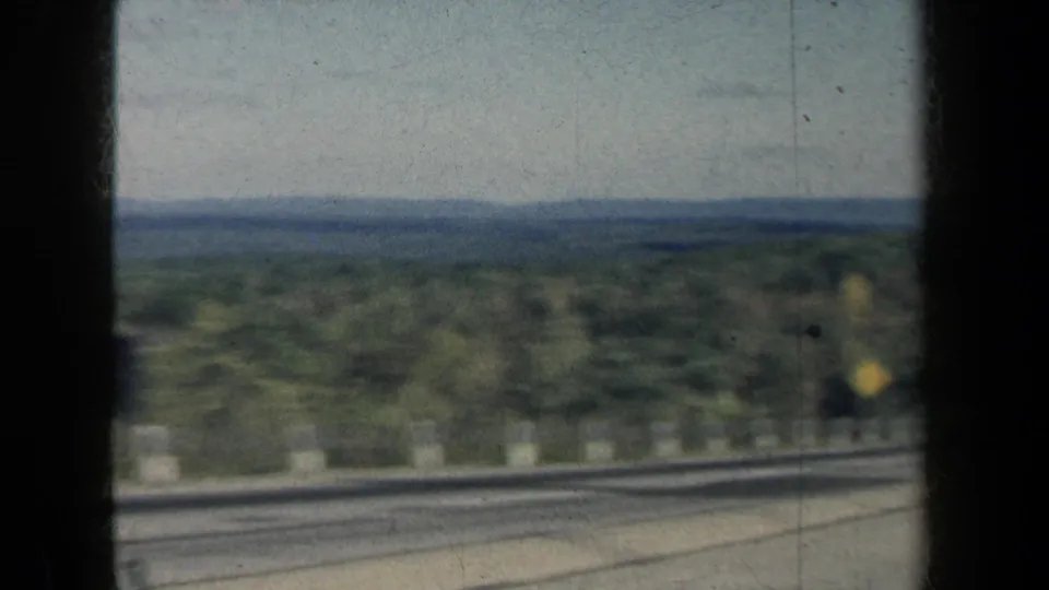 Motion-blurred 8mm view from a moving vehicle across guardrails, trees, and distant blue water in New York.