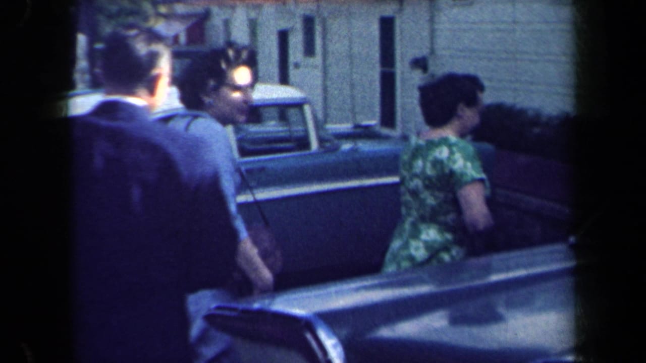 Women and a man stand near a parked car outside a house.
