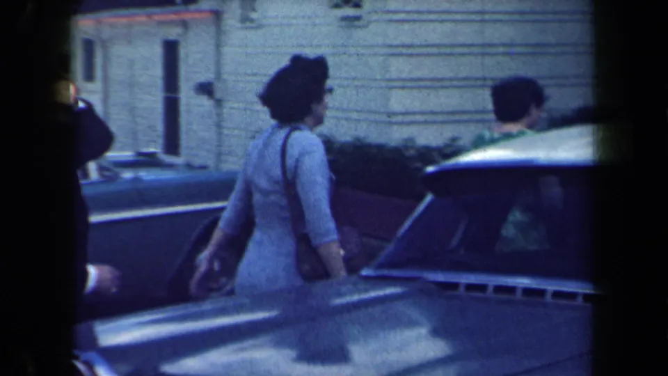1959 New York home-movie frame showing women in dresses walking beside parked cars near a house.