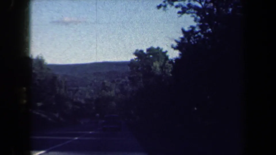 Rural New York road scene with roadside trees, a distant hill, and shadowed vegetation in 1959 8mm footage.