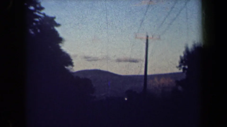 1959 8mm frame of a rural New York roadside with a wooden utility pole, rolling hill, trees, and soft evening-toned sky.