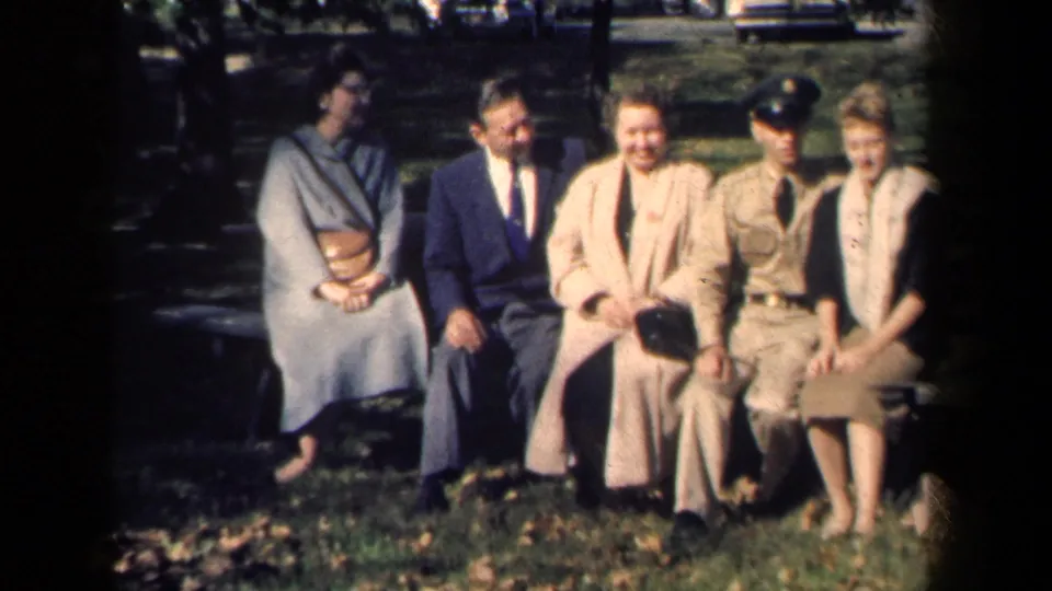 Five adults in late-1950s dress sit closely on an outdoor bench in New York, with grass, trees, and a parked car behind them.