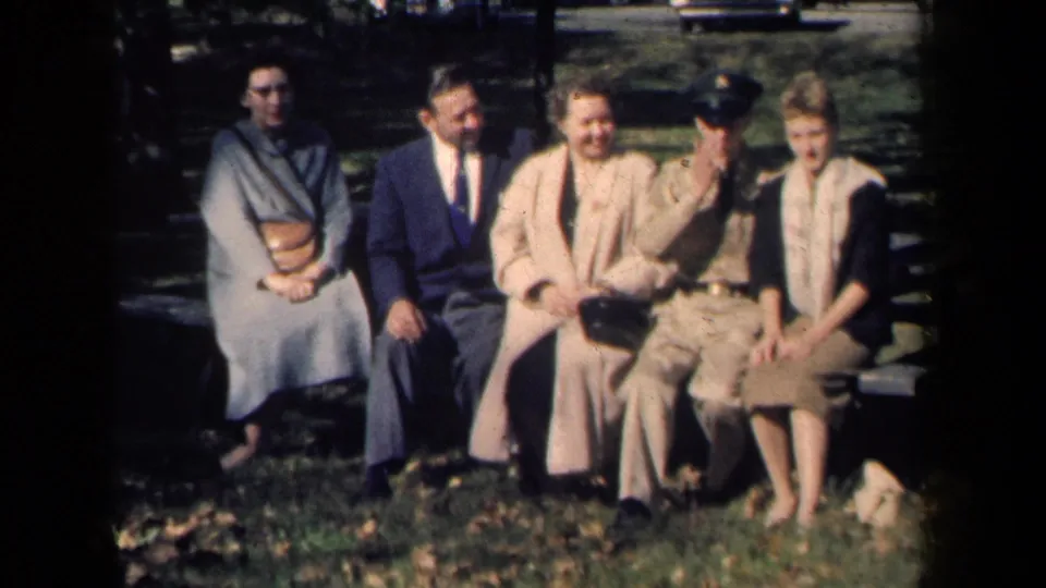 1959 New York home-movie frame of five adults smiling on a park bench, with one person in a uniform-like cap and trees behind them.