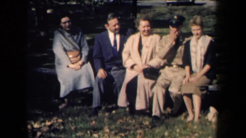 In 1959 New York home-movie footage, five people sit on a park bench with autumn leaves on the grass.