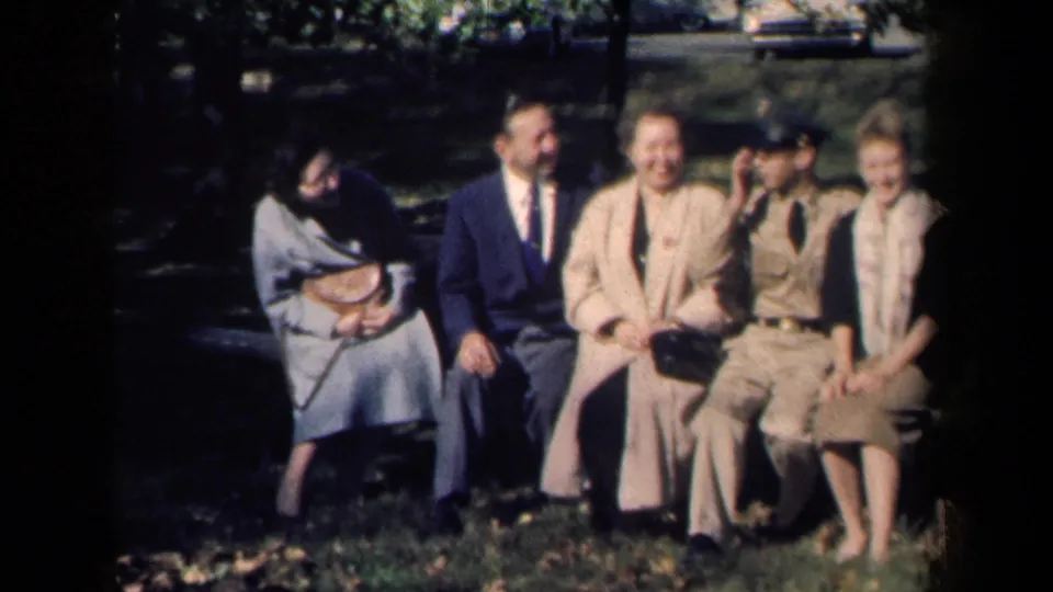 A 1959 New York park-bench group laughs together, with soft 8mm color and shaded grass in front.