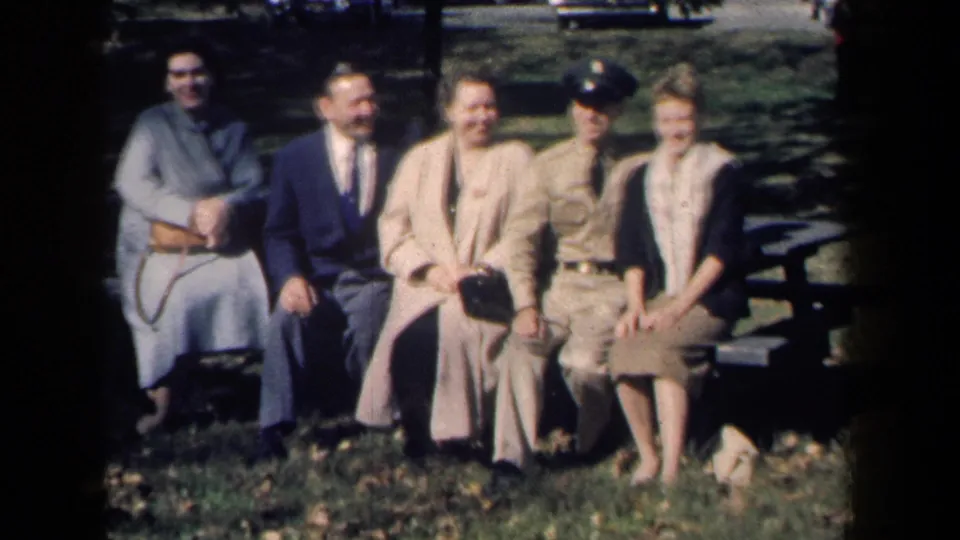 Five adults sit on a park bench in New York home-movie footage from 1959, with a uniformed man, coats, grass, and trees visible.