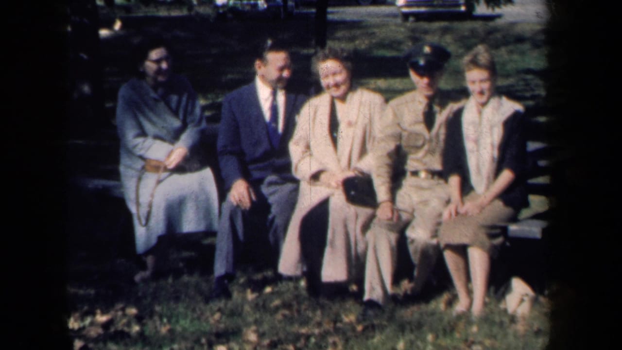 Five seated adults smile on a bench with grass, trees, and parked cars behind them.