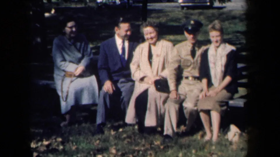 The seated group smiles toward the camera in 1959 New York park footage, with fallen leaves and parked cars in the background.