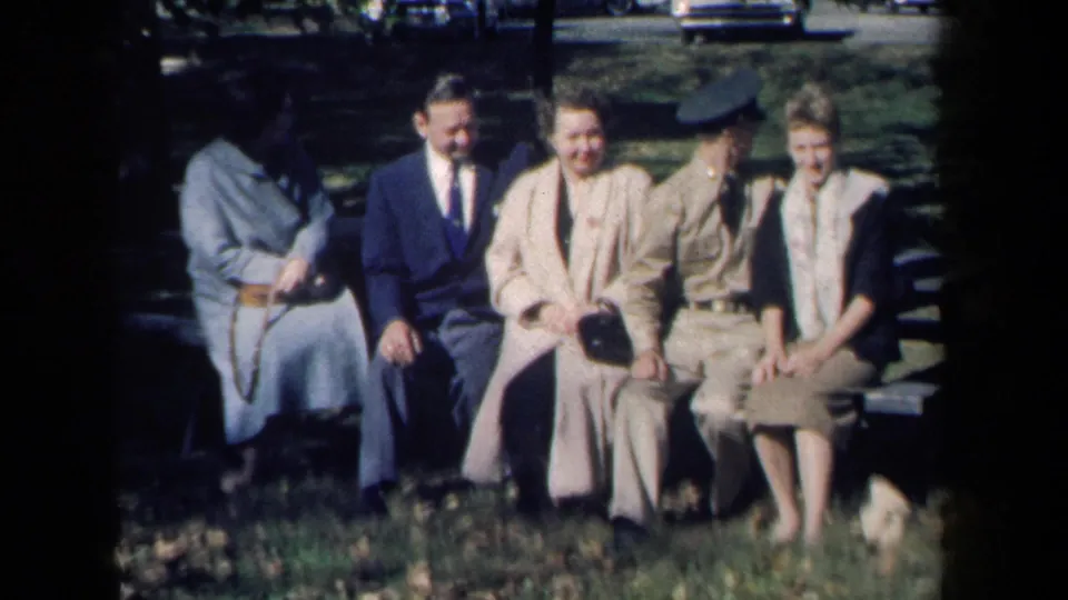 The group remains seated in the New York park scene, with the uniformed man turned slightly and the others smiling in soft 8mm color.