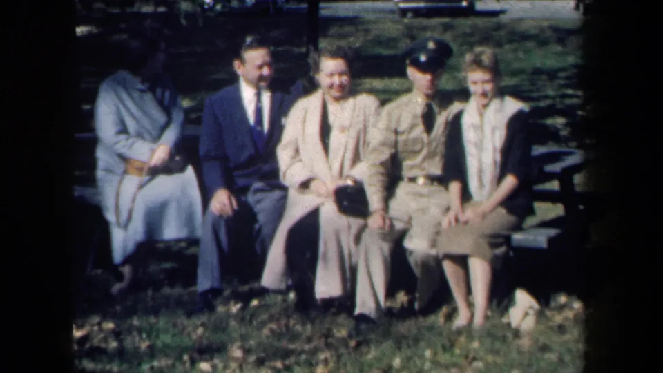 1959 New York home-movie frame showing five adults seated on a park bench, including a uniformed man, in a sunny outdoor setting.