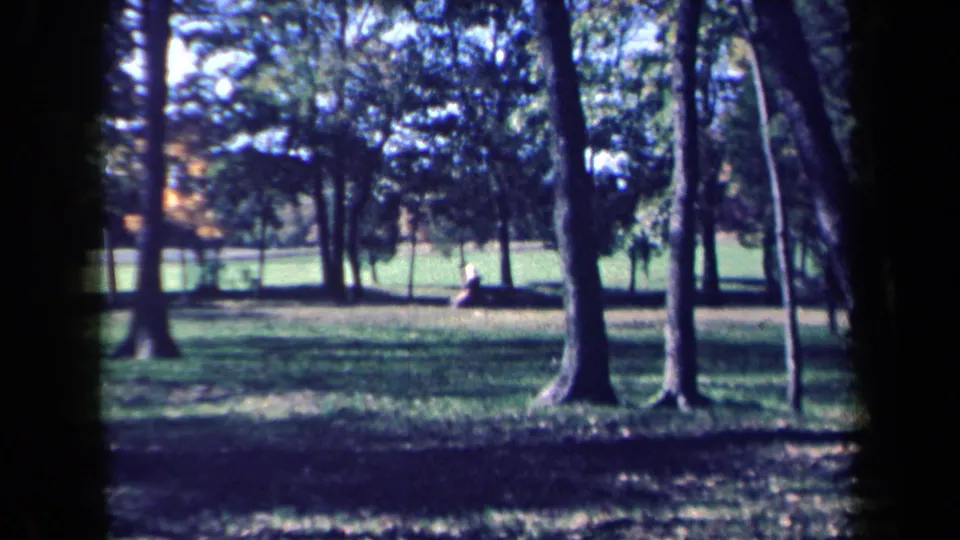 A 1959 New York home-movie frame shows shaded park trees, lawn, and a distant person seated by a bench.
