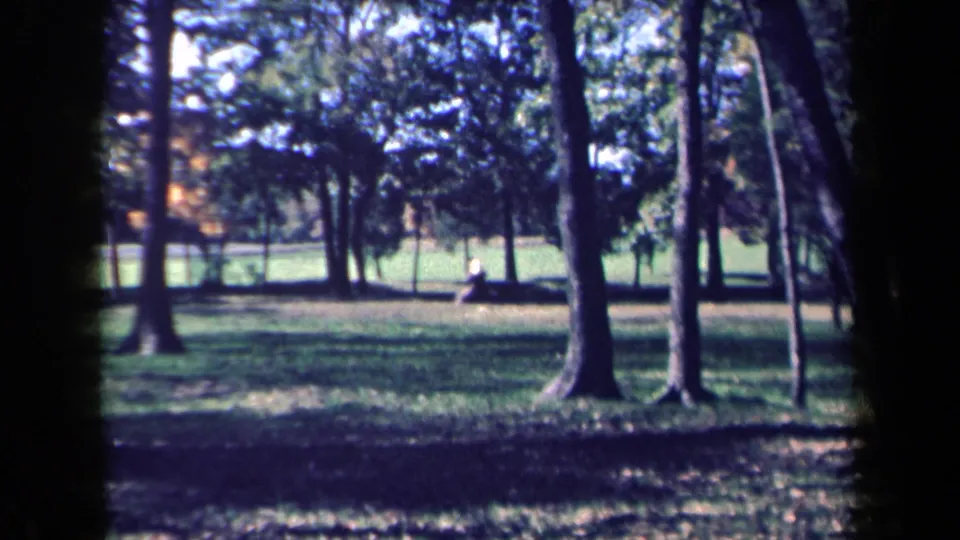 1959 New York home-movie frame showing a shaded grassy park with tall trees and a small seated figure in the distance.