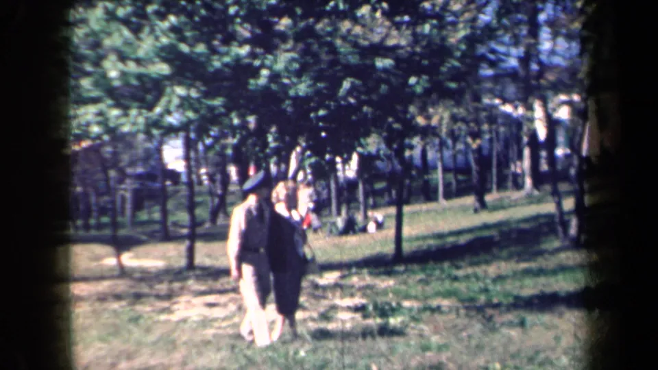 1959 New York home-movie frame showing an elderly couple walking through a grassy park under trees.