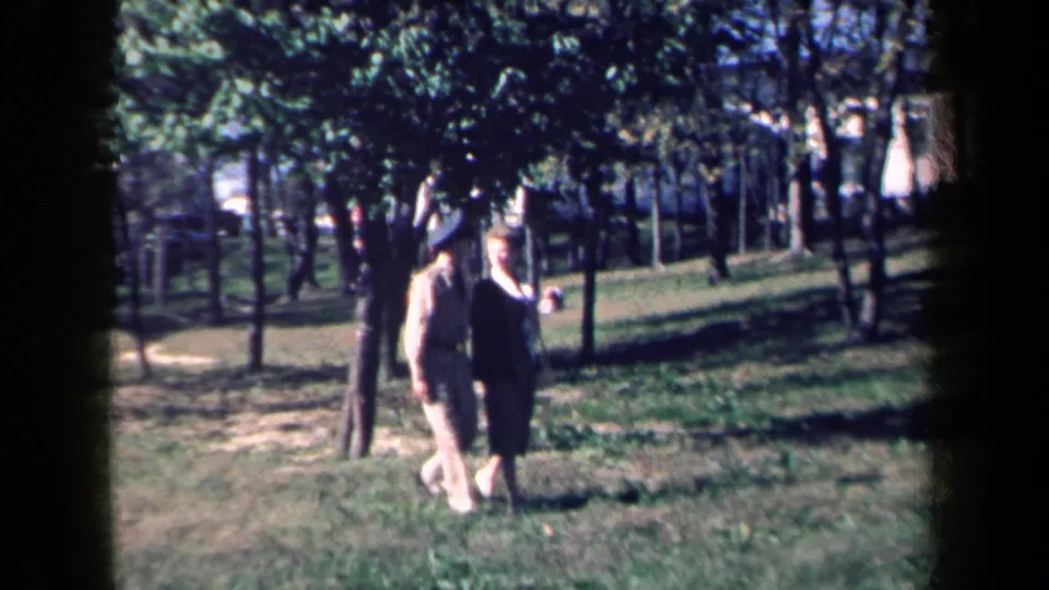 1959 New York home-movie frame of an elderly couple moving through grass in a tree-filled park setting.