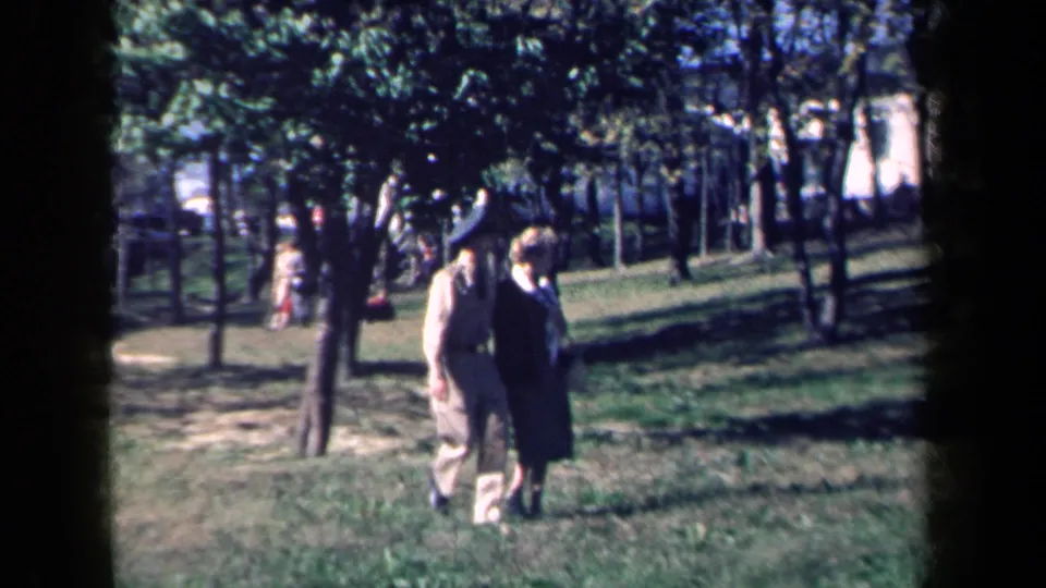 In 1959 New York home-movie footage, a couple walks arm in arm through a grassy park, with trees and distant visitors in the background.