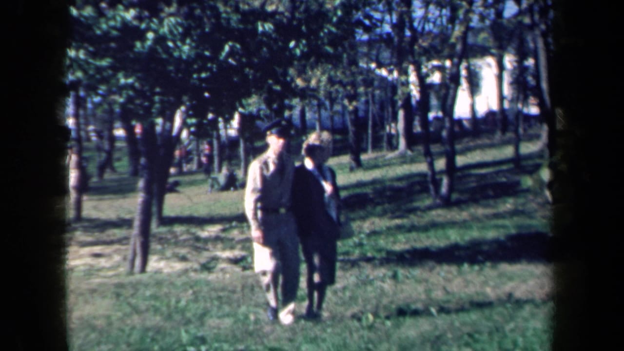 The couple moves closer through a New York park in 1959, the man in a light uniform-style outfit and the woman in dark clothing.