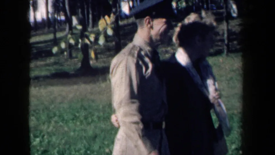 From the side, the man in uniform and woman continue their arm-in-arm walk through the sunlit park setting.
